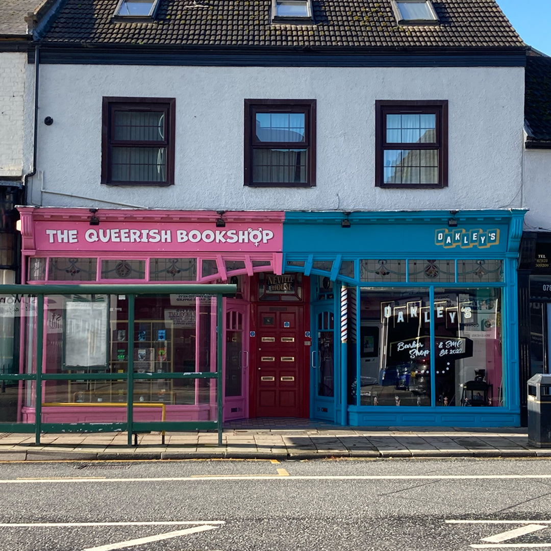 The Queerish Bookshop storefront is bright pink with the capitalised logo on the sign at the front. The pink shop stands next to a blue shop, Oakley's Barbers.
