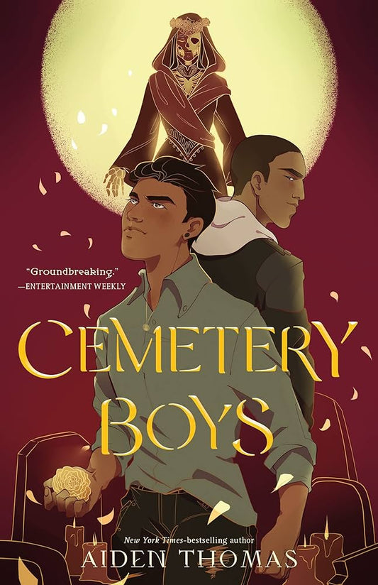 Two Latino teenagers stand back to back with tombstones behind them. A skeleton in a red robe hovers above them with the moon shining brightly behind her. The title "Cemetery Boys" sits in the centre.