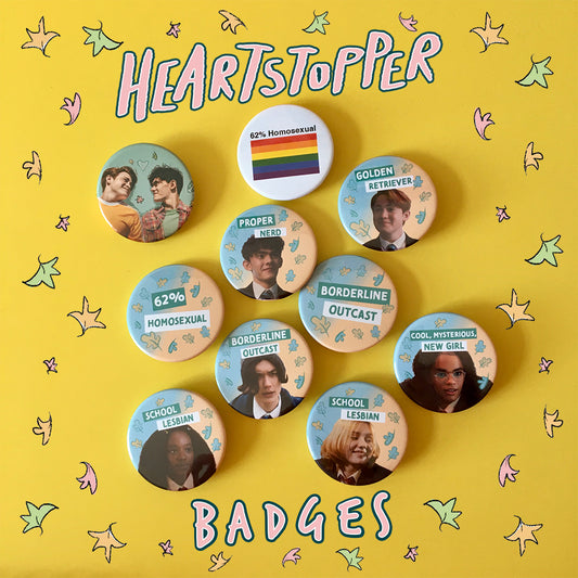 On a yellow desk are ten badges, all Heartstopper themed. Some have references from the show, like 62% Homosexual, but the majority of them have an image of a Heartstopper character on them. There is text that reads “Heartstopper Badges” and doodled leaves decorate around it. 