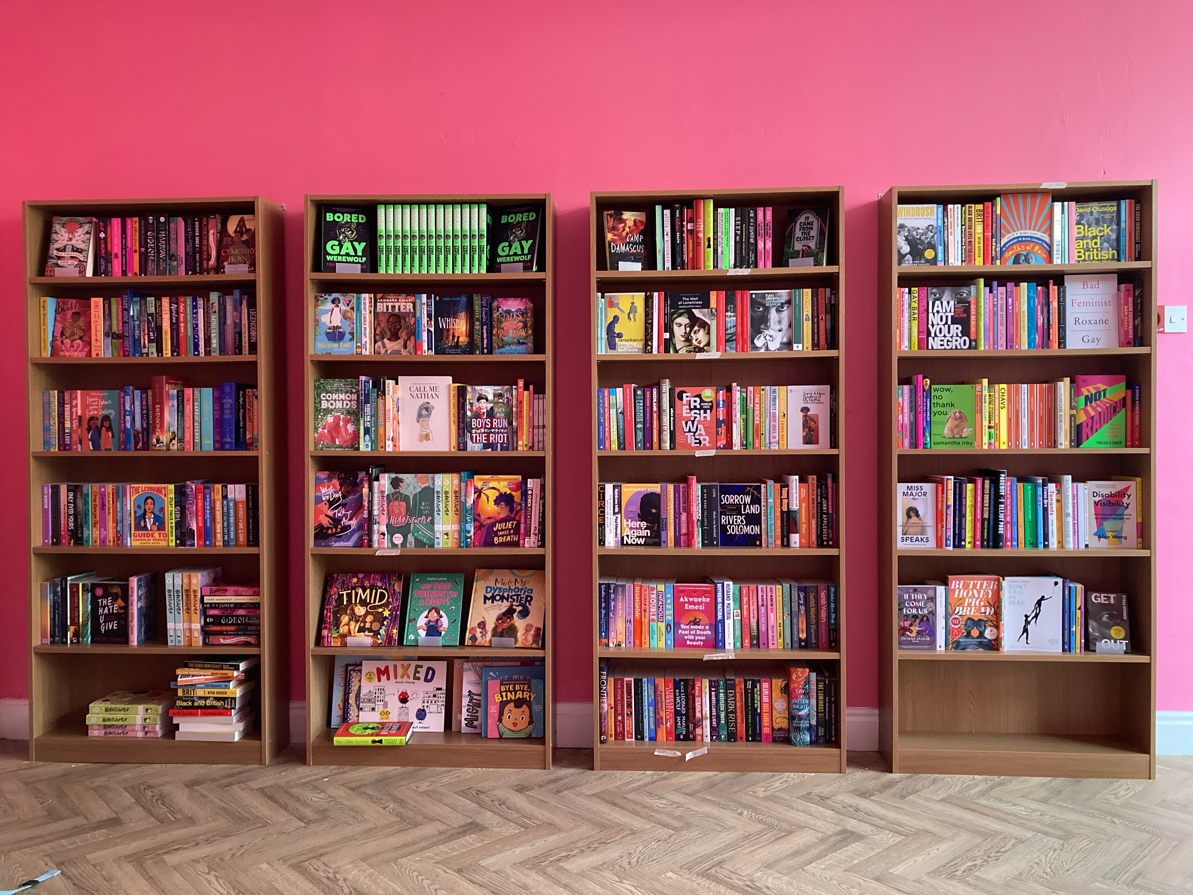 Four oak bookcases filled with queer and radical books stand against a bright pink wall.