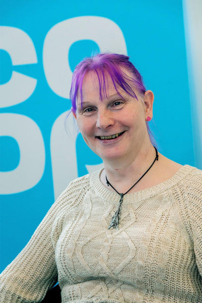 Emma Roebuck, a white trans woman with purple hair wearing a cream jumper, smiles at the camera.