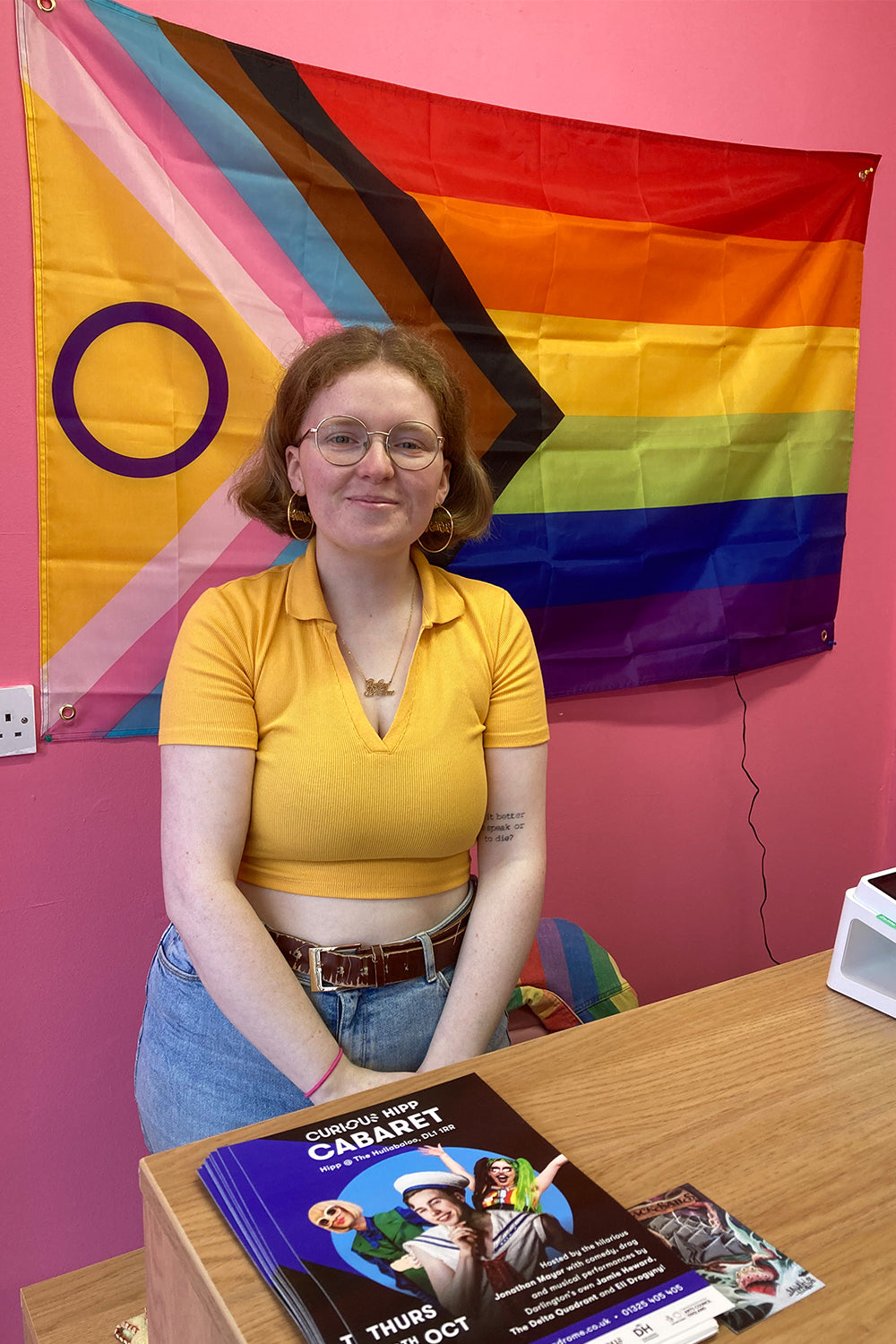 Megan Hemsley, a white queer woman with short gingery hair in a yellow cropped shirt and blue jeans, smiles at the camera while standing in front of a Pride flag.