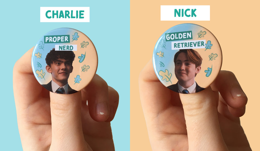 Nick and Charlie from Heartstopper are printed on two badges. Charlie is a white teen boy and has text that reads “Proper Nerd”. Nick is a white teen boy and his badge has text that reads “Golden Retriever.” 